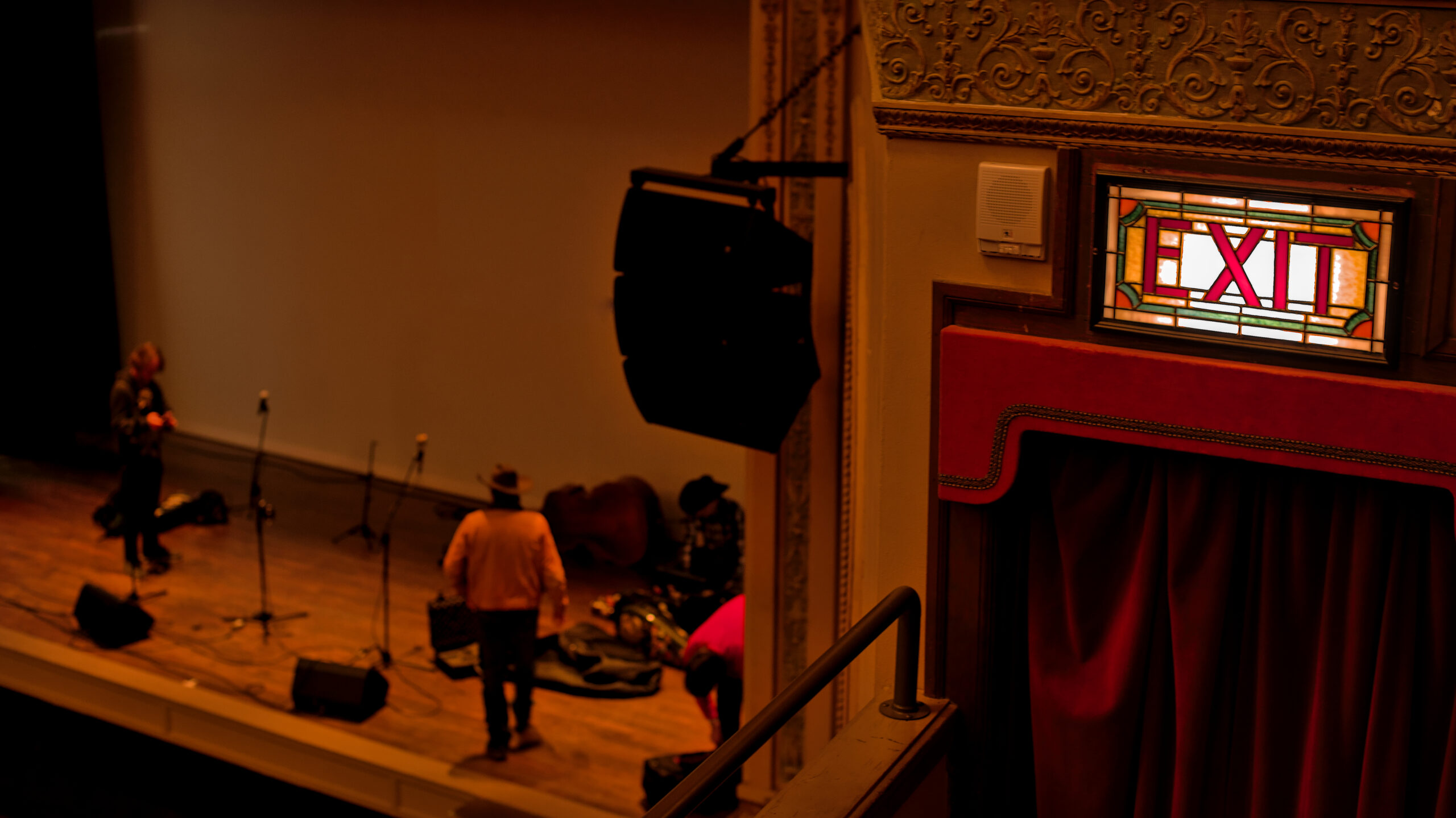 An exit sign lit up in the foreground while The Wardens uncase their instruments on the stage below at the Columbia Theatre, Copyright Jacob Greenslade